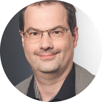 Eine Person mit Brille und grauer Jacke über einem dunklen Oberteil. 