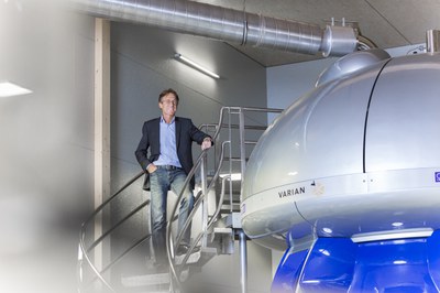 Prof. Dieter Willbold links auf einer Treppe stehend, rechts im Bild ein Ausschnitt eines 900-Mhz-NMR-Spektrometers, das in Willbolds Forschung zur Anwendung kommt.
