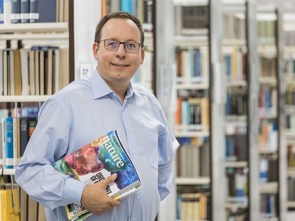 Bernhard Mittermaier mit Magazinen unter dem Arm in der Bibliothek stehend.