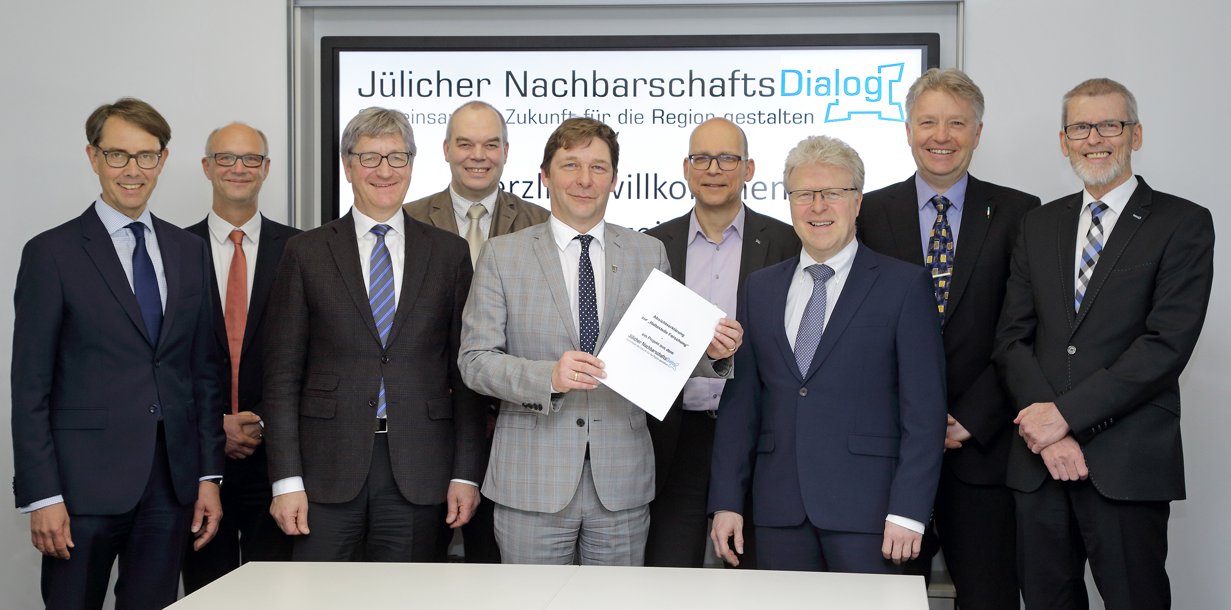 Gruppenbild der Unterzeichner vor einer Wand mit dem Logo des Jülicher Nachbarschaftsdialogs