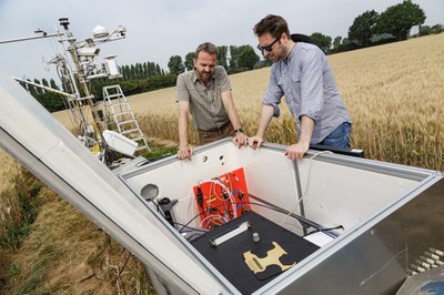 Zwei Wissenschaftler stehen auf einem Kornfeld mit gelbem Getreide vor einem großen Container mit Messgeräten