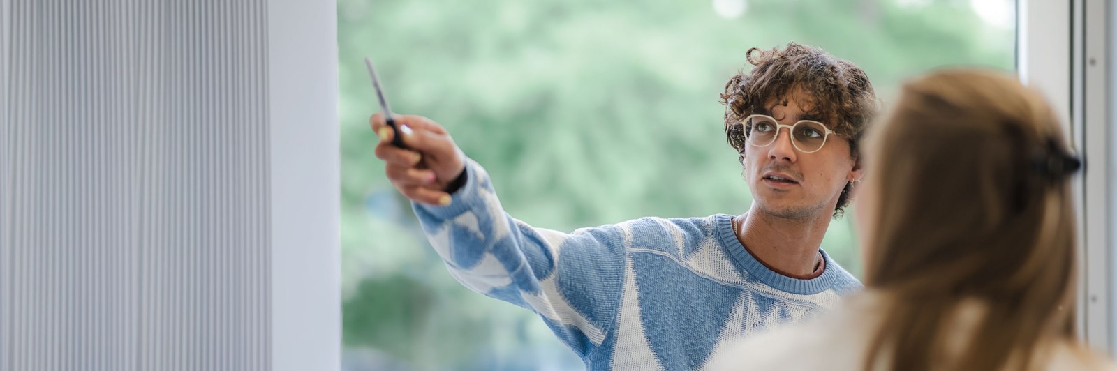 Eine Person mit Brille in einem karierten Oberteil hält einen Gegenstand in der Hand, während eine andere Person mit langem Haar von hinten sichtbar ist. (Mistral: Mistral Medium 3.5, 2026-06-24)