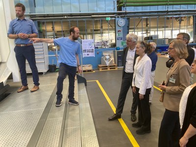 Mehrere Personen stehen in einer Halle mit industrieller Umgebung, während eine Person auf ein Gerät zeigt und eine andere Person gestikuliert.