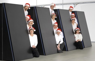 Mehrere Personen in weißer Kleidung und roten Mützen stehen hinter großen, geneigten, dunklen Flächen in einem hellen Raum.