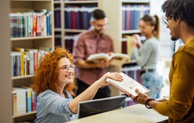 Mehrere Personen in einem Raum mit Regalen voller Bücher, eine Person reicht ein Buch an eine andere, während im Hintergrund weitere Personen Bücher betrachten. (Mistral: Pixtral Large 2411, 2026-03-17)