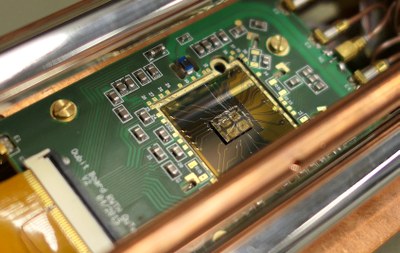 Eine grüne Platine mit mehreren elektronischen Bauteilen und Kupferkontakten in einem metallischen Gehäuse. (Mistral: Pixtral Large 2411, 2026-03-04)