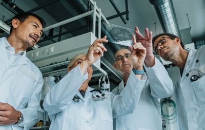 Mehrere Personen in weißen Kitteln untersuchen gemeinsam ein durchsichtiges Objekt in einem Raum mit industriellen Geräten im Hintergrund. (Mistral: Pixtral Large 2411, 2026-03-17)