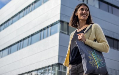 Eine lächelnde Person mit langen Haaren steht vor einem modernen Gebäude und hält eine Tasche mit buntem Muster. (Mistral: Pixtral Large 2411, 2026-03-04)