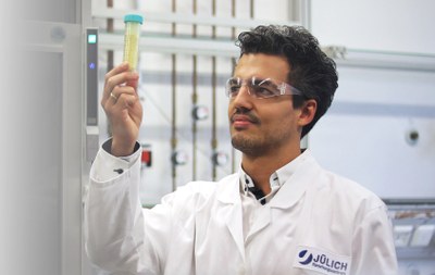 Eine Person in Schutzbrille und Laborkittel hält ein Reagenzglas mit einer Flüssigkeit vor einem Gerät.