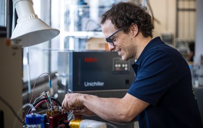 Eine Person lächelt, während sie an einem Gerät mit Kabeln und Schaltern arbeitet, neben einer Lampe und einem Gerät mit Knöpfen. (Mistral: Pixtral Large 2411, 2026-03-05)