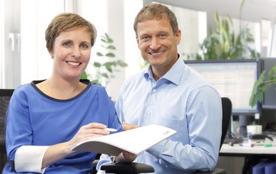 Zwei lächelnde Personen sitzen nebeneinander in einem Büro, eine hält ein Blatt Papier, im Hintergrund sind Pflanzen und ein Gerät sichtbar.