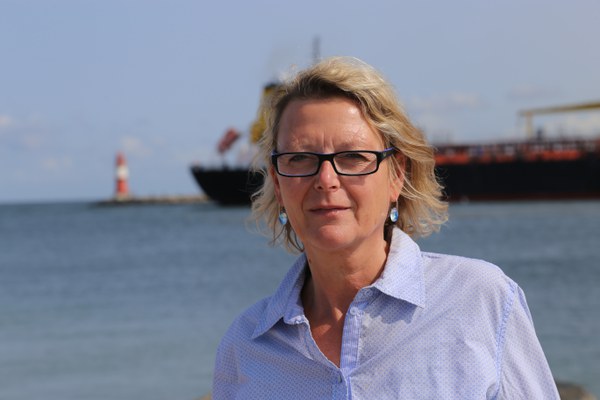 Frau Fretzdorff lächelt im Portrait freundlich in die Kamera. Im Hintergrund befindet sich der Molenausgang von Warnemünde mit einem herausfahrenden Containerschiff und einem roten Leuchtturm.