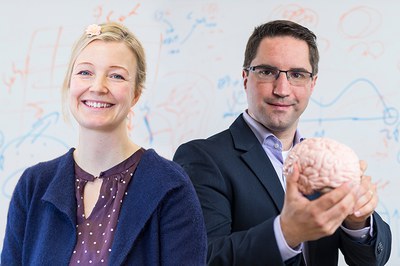 Eine Mitarbeiterin und ein Mitarbeiter stehen vor einem Whiteboard. Die Mitarbeiterin lächelt. Der Mitarbeiter lächelt leicht und hält ein Modell eines Gehirns in den Händen.