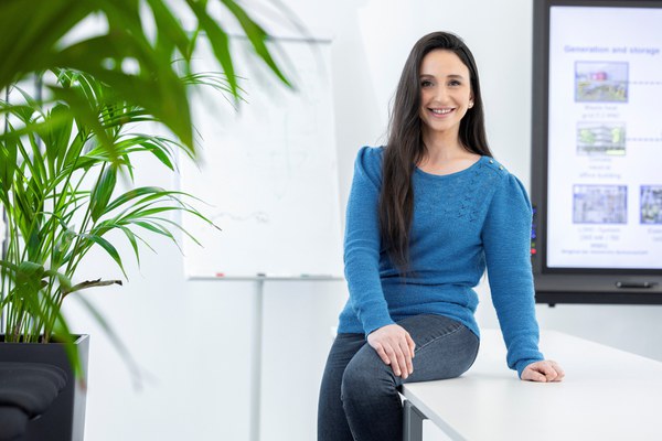 Eine Kollegin lehnt an einem Tisch und lächelt freundlich in die Kamera. Im Hintergrund ist ein Bildschirm mit einer wissenschaftlichen Präsentation zu erkennen.