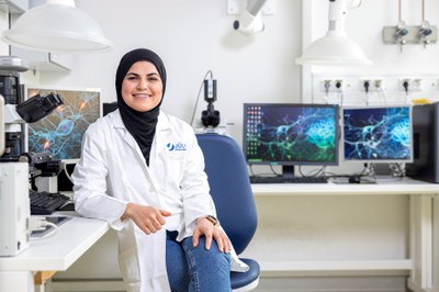 Eine Postdoc-Kollegin sitzt auf einem Stuhl in einem Labor. Sie trägt einen Kittel und im Hintergrund sind Computerbildschirme zu sehen.