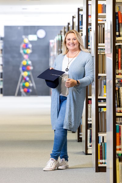 Eine Mitarbeiterin steht an ein Bücherregal geleht da und lächelt in die Kamera. Sie hält einen Graduationshut in den Händen.