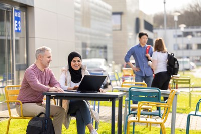 Vier Personen befinden sich auf dem Campus des Forschungszentrums. Zwei sitzen an einem Tisch und schauen gemeinsam auf einen Laptop. Im Hintergrund stehen zwei weitere Personen, die sich unterhalten.