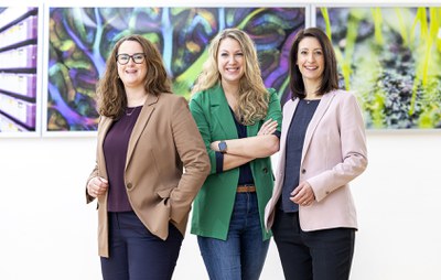 Drei Frauen stehen nebeneinander und lächeln in die Kamera. Im Hintergrund hängen bunte Bilder an einer Wand.