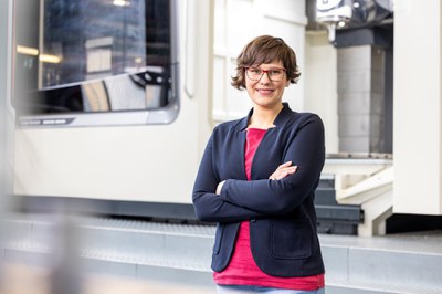 Portrait einer jüngeren Mitarbeiterin in moderner Businesskleidung, die enthusiastisch in die Kamera lächelt, vor dem Hintergrund einer Großmaschine in der Werkstatt-Infrastruktur