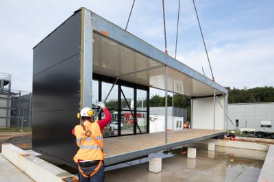 Ein großer Container wird von einem Kran gehoben, während eine Person in Schutzkleidung und Helm daneben steht. (Mistral: Pixtral Large 2411, 2026-03-05)