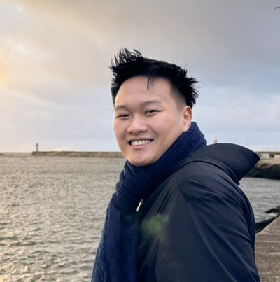 Eine Person mit dunklem, spitzem Haar und einem blauen Schal steht vor einem Gewässer mit einer Pier und zwei Leuchttürmen im Hintergrund unter bewölktem Himmel. (Mistral: Mistral Medium 3.5, 2026-09-18)