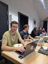 Mehrere Personen sitzen in einem Raum an Tischen und arbeiten mit Geräten, während eine Person in den Vordergrund blickt. (Mistral: Pixtral Large 2411, 2026-03-17)