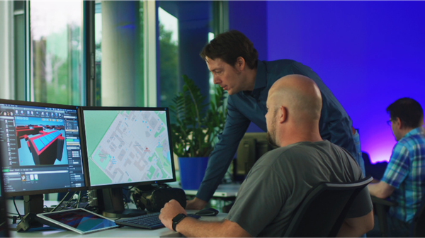Employees sit at their desks in front of their screens in the office. Some are talking to each other.