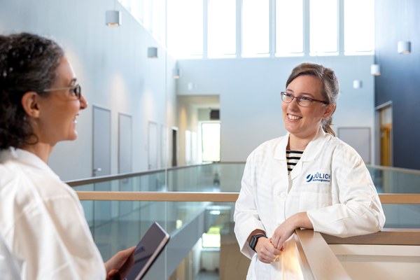 Two women in white lab coats are talking in a modern, bright building with large windows.