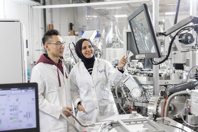 Two people in white lab coats stand in a laboratory in front of technical equipment and a monitor, one person points at the screen
