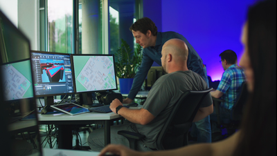 Several people sit at desks with multiple monitors displaying technical drawings