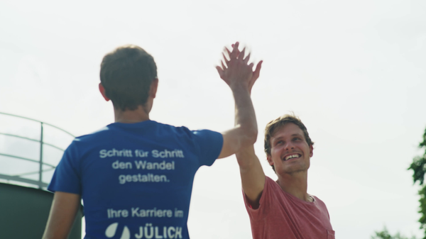Two young men give each other a high-five outdoors. One is wearing a blue T-shirt with the words ‘Shaping change step by step. Your career at Jülich’, the other wears a red T-shirt and smiles. The scene conveys team spirit and joy.