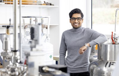 A scientist leans against a large vessel in a laboratory and smiles at the camera