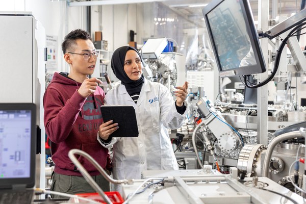 A young man in a hoodie and a young woman with a headscarf and white coat are standing in the centre of a technical system. They are looking at a digital screen.
