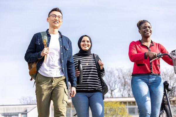 Three young international colleagues are walking across the Jülich campus together. He has a rucksack over his shoulder and one of the women is pushing a bicycle.
