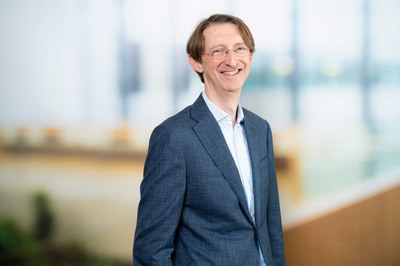A person with short hair and glasses, wearing a suit and white shirt, smiling and standing in a modern indoor setting with glass walls and natural light. (Mistral: Pixtral Large 2411, 2026-03-17)