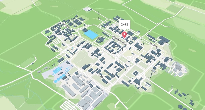 Aerial view of a campus with multiple buildings, green spaces, pathways, and a central area marked with a red pin. (Mistral: Pixtral Large 2411, 2026-05-12)