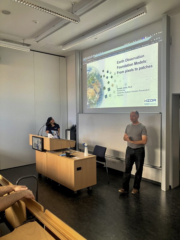 Two people stand in a room with chairs, one near a table with a laptop and a bottle, the other near a large screen displaying an image. (Mistral: Mistral Medium 3.5, 2026-09-07)