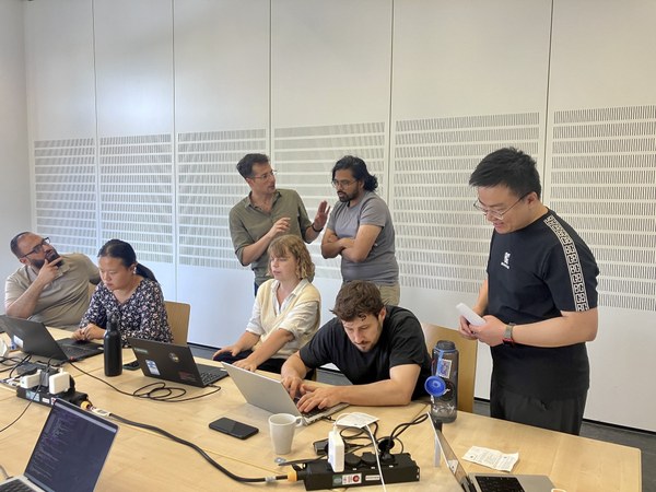 Several people are gathered around a table with laptops, a small device, and various objects like bottles and cups. (Mistral: Mistral Medium 3.5, 2026-09-07)
