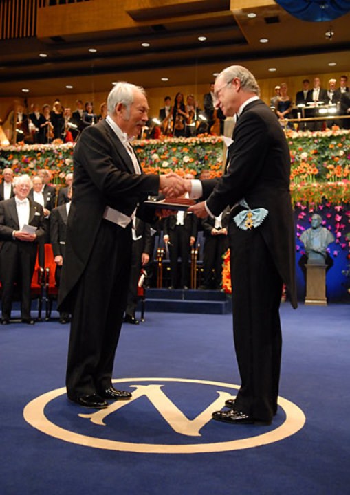 Two men in formal attire shake hands on a stage with a large audience seated in the background. (Mistral: Pixtral Large 2411, 2026-03-04)